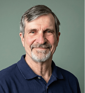 A person with short graying hair and a beard is smiling gently while wearing a dark blue collared shirt against a plain gray background. Description generated by AI